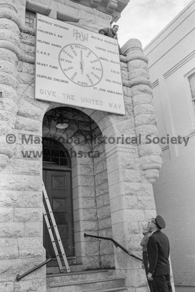 Photographer: Tashiro
October 16, 1974
United Way
United Way campaign rolling smoothly
PPUW clock
This clock will record the progress of the 1974 Portage Plains United Way campaign receipts. It was built by Portage firemen and installed Wednesday in front of city hall with assistance of members of the city works department. Fire Chief Harold Bradon and United Way director Mrs. Sharon Bruinooge look up at the clock while city works department foreman Fred Dell glances down after fastening the clock in place over the door of city hall. VP-2-00265-004.jpg
