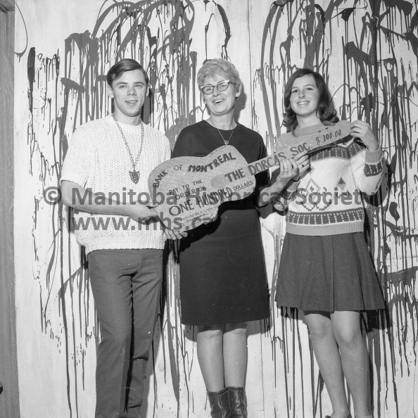 Photographer: Tashiro
January 3, 1969
Dorcas Soc.
Dorcas ladies receive $100 from Burlap Den coffee house
The Dorcas Society, which earlier this week discovered its 1968 campaign for funds trickled over the top, received a healthy boost Friday. Folk Episode ‘68, a Boxing Day concert performed by artists from the Burlap Den coffee house, netted the society $100. Making the presentation of the cheque — appropriately in the form of a guitar — to Mrs. Lois Forsythe of the Dorcas Society are Rick Poltaruk and Linda Sanderson. VP-1-00726-001.jpg