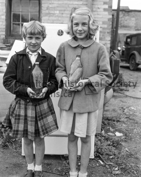 Photographer: Unknown
September 24, 1952
News
Louise and Joyce Shyiak - Cucumber grown in bottle
Cucumbers in a bottle — Ships in bottles well that's something different, and something that is seen quite often. But cucumbers in a bottle? These two little misses, Louise and Joyce Shyiak aged nine and 10, decided they'd try something different. They put the cucumber vine into the two vinegar bottles and waited for results. Here they are holding their prize possessions fully — grown cucumbers in bottles. One bottle contains two cucumbers fully grown. VP-1-04407-001.jpg